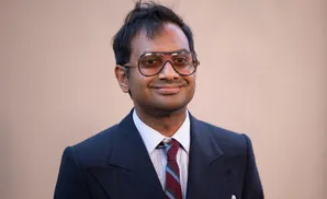 A man wearing a suit and aviator glasses smiles at the camera.