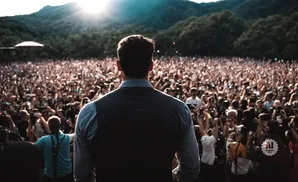 Man in a vest facing a large crowd with hands raised, a sunny, mountainous background.
