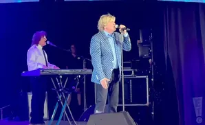A male singer in a blue polka-dot jacket stands on stage with a microphone, next to another man playing a keyboard.