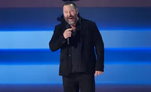 Comedian Bert Kreischer laughs while holding a microphone on a stage with a blue background.