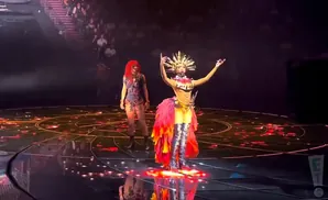 A performer in a flamboyant costume with a golden headdress and feathered skirt poses on a stage.
