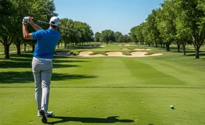 Golfer in blue shirt swings club on a green golf course with sand traps and trees.