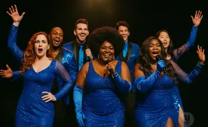 A group of seven singers in dazzling blue sequined outfits perform onstage, with two women in the foreground holding microphones.