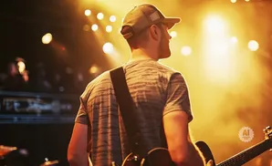 A guitarist in a cap and striped shirt performs on a brightly lit stage, holding his guitar.