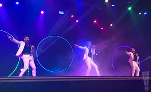 Three men in white suits sing on a stage with purple and blue lights and glowing circles.