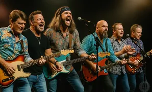 A band of seven men play guitars and sing on a stage with a dark background.