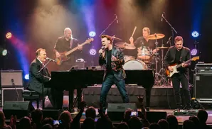 A band performs on stage with a pianist in a suit, a singer in a floral jacket, a guitarist, and a drummer.