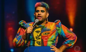 A person with rainbow-colored hair, wearing a colorful, abstract-patterned top, speaks into a microphone on a stage.