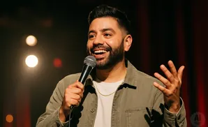 A smiling man with a beard holds a microphone and gestures with his hand while on stage.