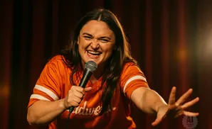 A woman in an orange shirt laughs while holding a microphone on a stage.