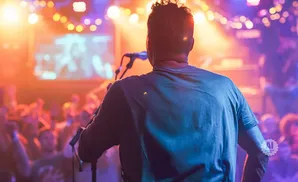 A man faces a cheering crowd at a concert, bathed in colorful stage lights.