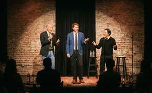Three men on a stage with brick backdrop, two holding microphones, performing for an audience.