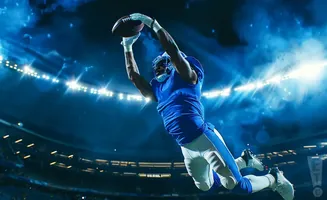 An American football player in a blue uniform leaps to catch a football under stadium lights.