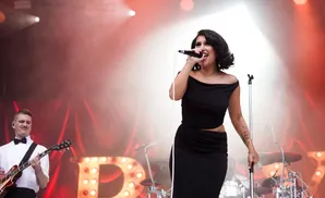 A female singer performs on stage in a black off-the-shoulder dress, with a guitarist in a white shirt playing in the background.