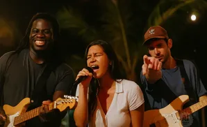 A band performs on stage. A Black man on the left plays guitar, a woman in the middle sings into a microphone, and a man on the right points at the camera.