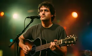 A man plays an acoustic guitar and sings into a microphone on a dimly lit stage with colorful lights.