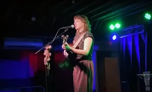 A person with shoulder-length hair plays an acoustic guitar and sings into a microphone on a dimly lit stage.