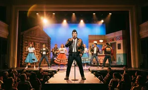 A musical performance on stage with singers and dancers in 1950s attire, with an audience watching.