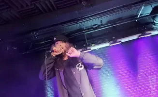 A rapper with long, curly hair and a baseball cap holds a microphone and performs on stage with purple and blue lights.