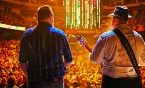 Two musicians on stage with a cheering crowd and colorful lights behind them.
