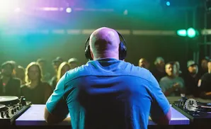 A bald DJ with headphones faces a crowd, with colorful lights illuminating the scene.