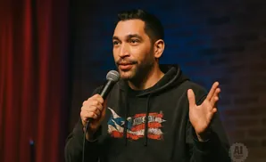 Comedian holding a microphone, wearing a dark hoodie with a distressed Puerto Rican flag design, gesturing with his free hand.
