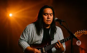 A person plays a guitar and sings into a microphone under warm stage lighting.