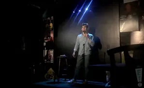 A man in a grey shirt and pants stands on stage holding a microphone and smiling.