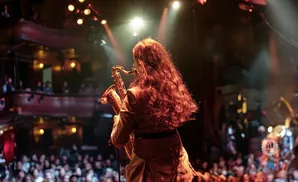 A musician with long, wavy red hair plays a saxophone on a dimly lit stage in front of an audience.