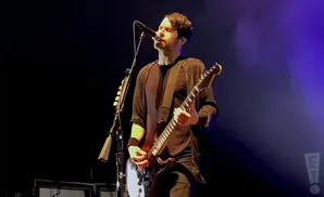 Musician playing guitar and singing into a microphone on a dimly lit stage with purple lighting.