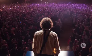 A person with curly hair stands facing a large, cheering crowd at a concert, lit by purple stage lights.