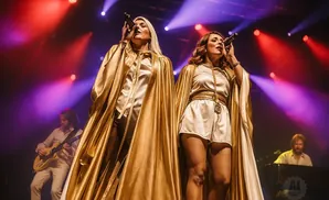 Two women in gold cloaks sing into microphones on stage, with a guitarist and pianist in the background.