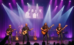 A band performs on stage under purple and white lights, with a photo of the band displayed on a screen behind them.