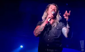 A tattooed man with long blonde hair and a mustache sings into a microphone on a dark, blue-lit stage.