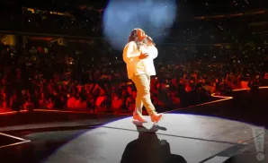 A person in a white shirt and tan pants sings on stage in front of a cheering audience.