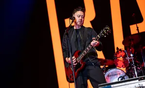A man in a black jacket plays a red electric guitar on stage with a large orange 