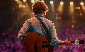 Musician playing an acoustic guitar on stage with blurred lights and audience in the background.