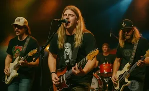 A band performs on stage with guitars and drums under stage lights.