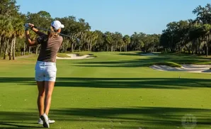 Golfer swings club on a sunny golf course with palm trees and sand traps.