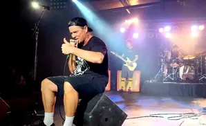 A man in a black cap and t-shirt sits on a speaker, holding a microphone and giving a thumbs-up.