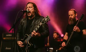 A rock band performs on stage with pink and purple stage lighting. The lead singer plays guitar and sings into a microphone.