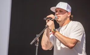 Man in white cap and t-shirt singing into a microphone with a gold watch on his wrist.