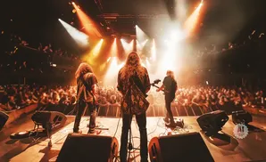 A band with long hair plays guitars on a stage in front of a cheering crowd.