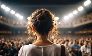 Woman with hair in a bun and gold jewelry faces a large audience, bathed in stage lights.