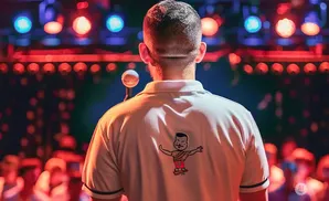 Man in a polo shirt with a cartoon logo, facing a stage with colorful lights and a microphone.