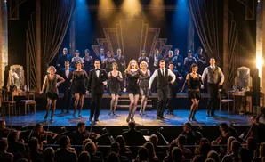 Stage performers in flapper dresses and suits dance and sing in a brightly lit musical production.