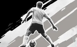 A black and white image of a soccer player from behind, about to kick a ball.