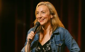 A blonde woman in a denim jacket holds a microphone and smiles