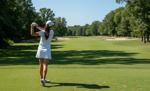 Golfer swings club on a sunny day on a green golf course with trees and sand traps.