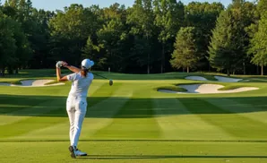 Golfer in white swings a club on a green golf course with sand traps and trees.